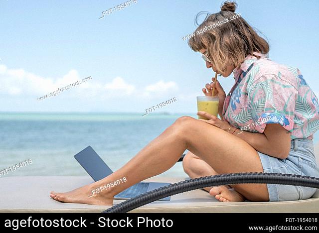 Woman sipping cocktail at laptop on beach lounge chair