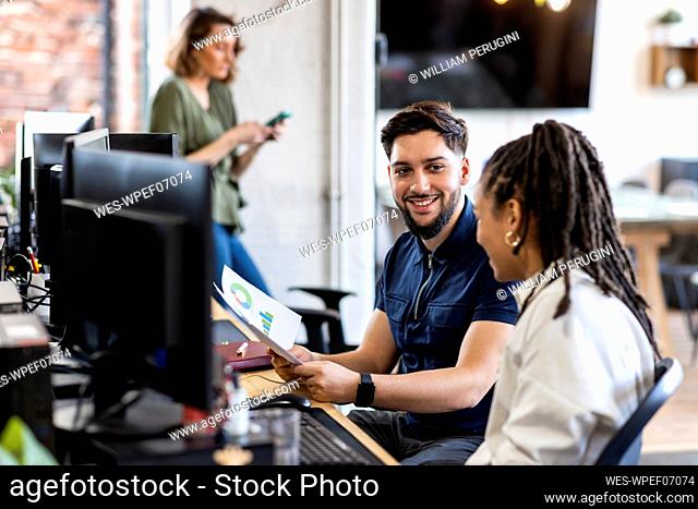 Happy businessman holding charts and graphs with colleague in front of computer