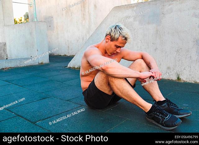 Young caucasian man sitting on work out ground and resting after training outdoors. Shirtless athletic man