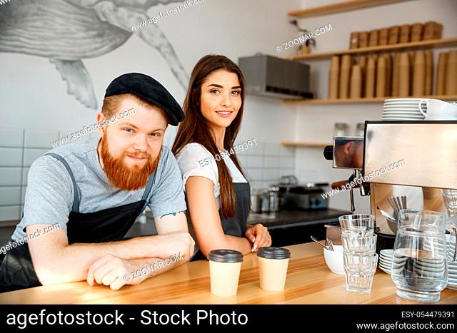 Coffee Business Concept - Positive young bearded man and beautiful attractive lady barista couple in apron looking at camera while standing at bar Couter ready...