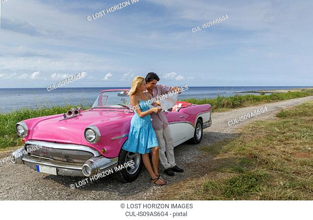 Young couple looking at map beside vintage convertible at coast, Havana, Cuba