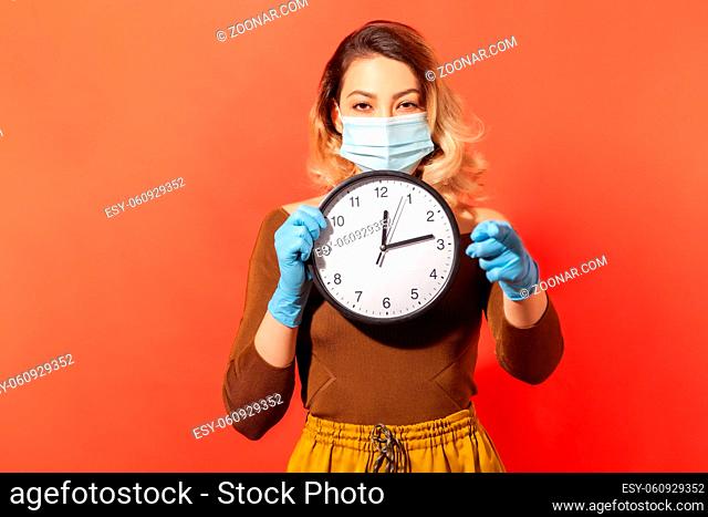 Time to stay home. Woman in surgical mask and gloves pointing to camera and holding clock, meaning quarantine, self-isolation while contagious disease spread