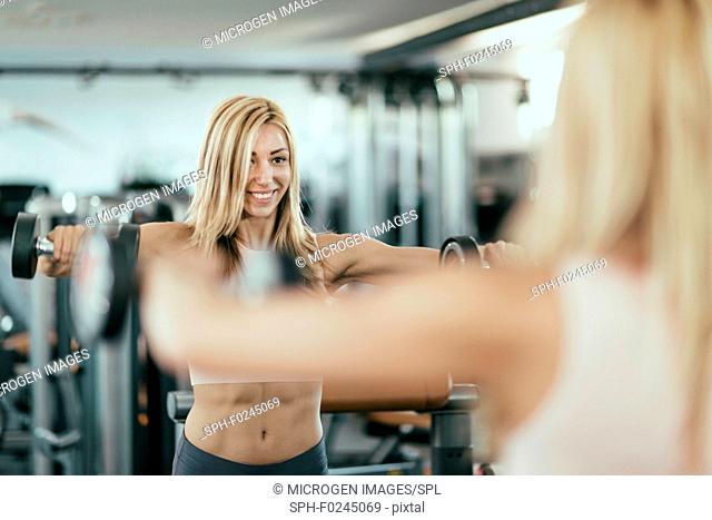 Young woman exercising with weights in front of the mirror in gym. Focus set on her face in the mirror