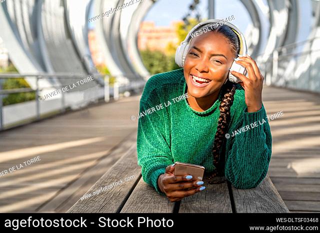 Happy woman listening to music through headphones and holding mobile phone lying on bridge