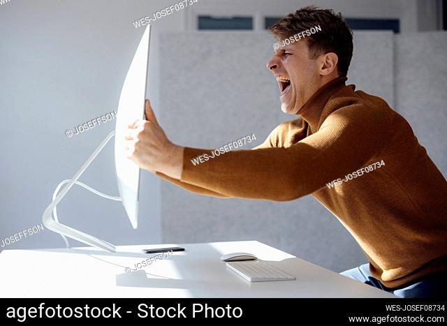 Angry businessman lifting computer monitor screaming at desk in office