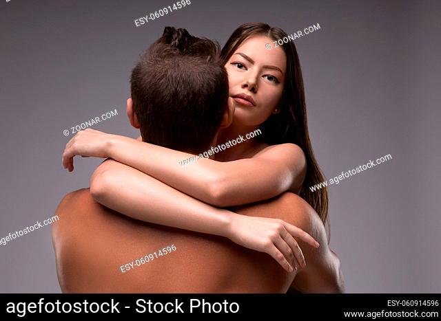 Young female model with long hair embracing shirtless male and looking at camera against gray background