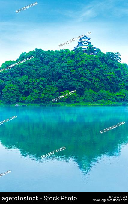 Inuyama Castle front facade from distance high above forest hill reflected in Kiso River water on blue sky day in Gifu Prefecture, Japan