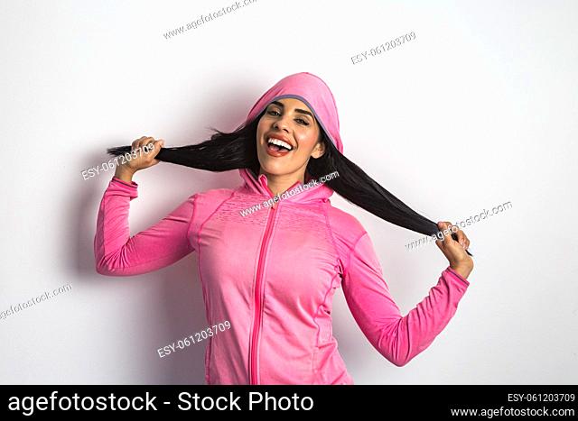 Joyful ethnic female model dressed in sports jacket touching hair and shouting happily in studio against white background while looking at camera