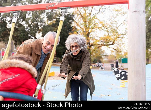 Playful grandparents pushing toddler grandson on swing at playground