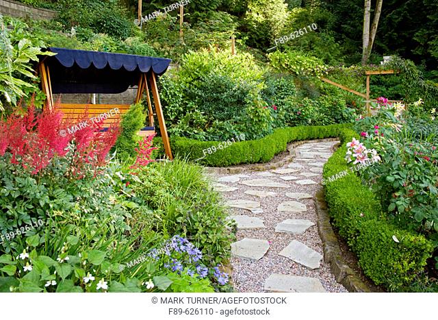 Covered wooden swing beside Red Astilbe w/ stepping stone path bordered by dwarf Boxwood hedges (Astilbe cv.; Buxus sempervirens)