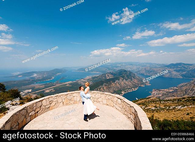 The groom holds the bride in his arms on the observation deck, a panoramic view of the Bay of Kotor behind of them . High quality photo