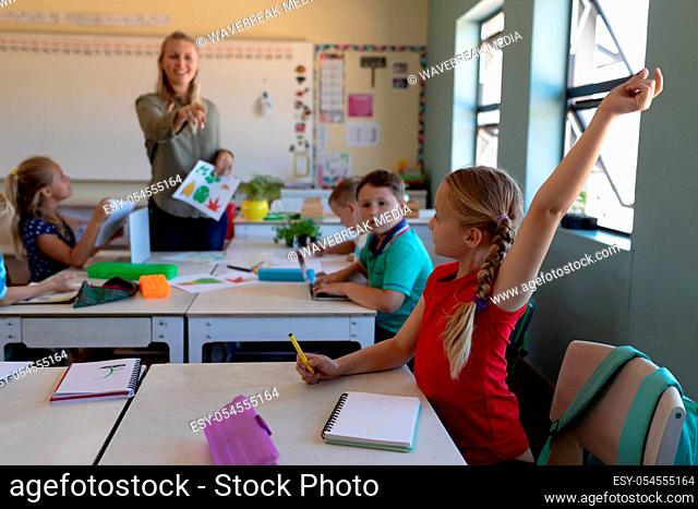 Side view of a Caucasian schoolgirl with blonde hair in plaits wearing a red t shirt sitting at a desk raising her hand during a lesson to answer a question in...