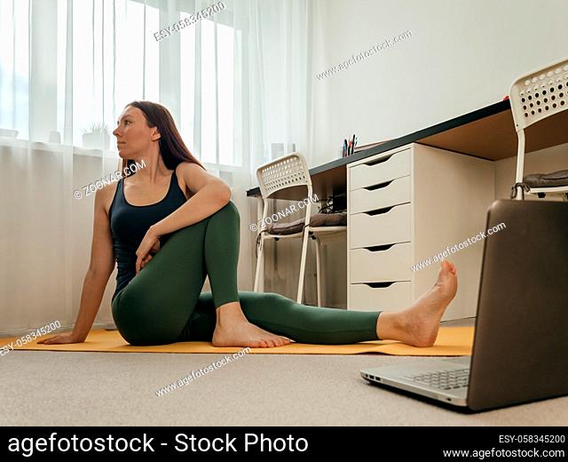 Young woman sitting on floor and doing yoga or stretching at home near laptop. Yoga online in small space home and wellbeing concept