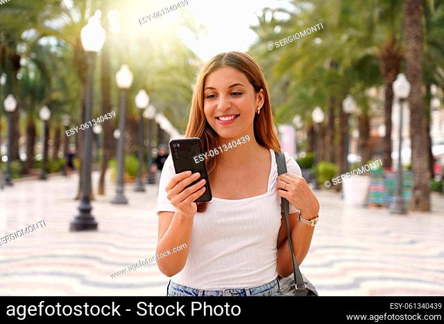 Medium shot young smiling woman uses smartphone reads news from social networks searches text sends mails online strolls at street