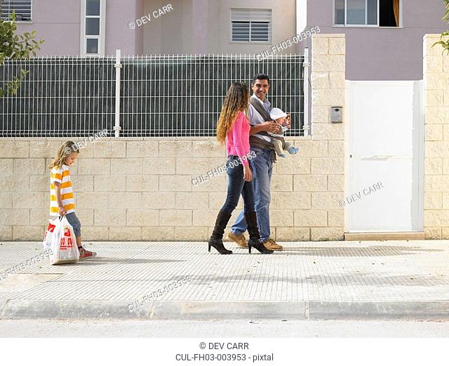Family walking on pavement - father with baby 5 months in sling , young daughter 4-6 with shopping dragging behind, Alicante, Spain