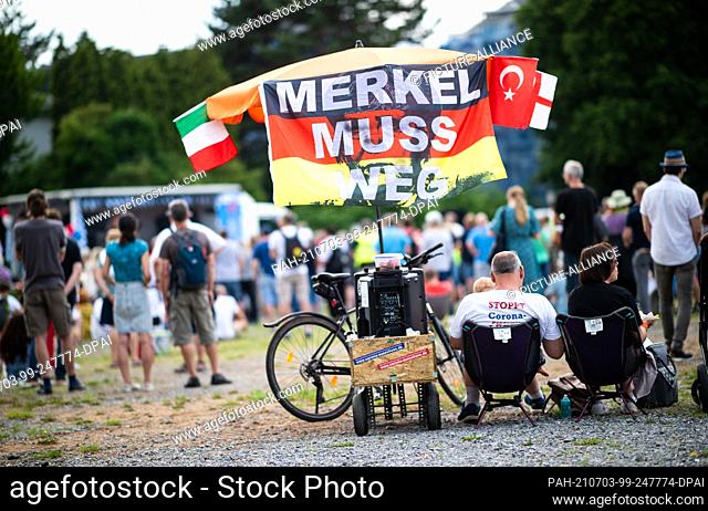 03 July 2021, North Rhine-Westphalia, Bochum: Two demonstrators sit on chairs next to an umbrella on which a poster with the inscription ""Merkel must go"" can...
