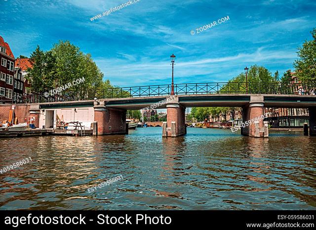 Bridge with iron balustrade on tree-lined canal, old buildings and sunny blue sky in Amsterdam. The city is famous for its huge cultural activity