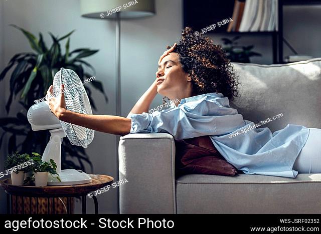 Young woman cooling off using electric fan lying on sofa at home