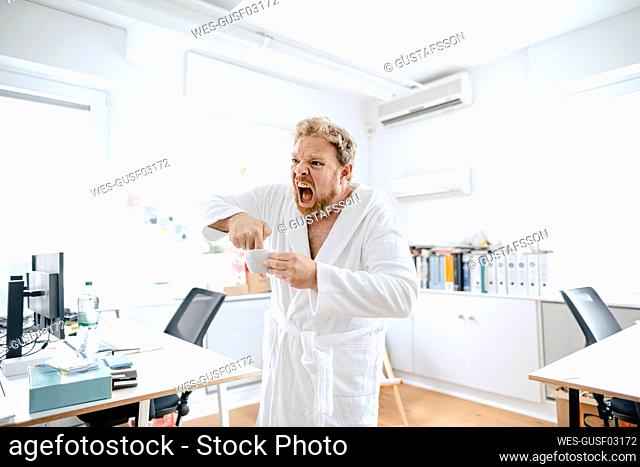 Screaming businessman wearing bathrobe in office putting his hand into coffee cup