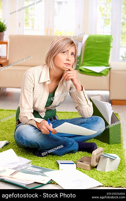 Worried woman thinking with paper in hand sitting on living room floor, checking documents, looking up, thinking