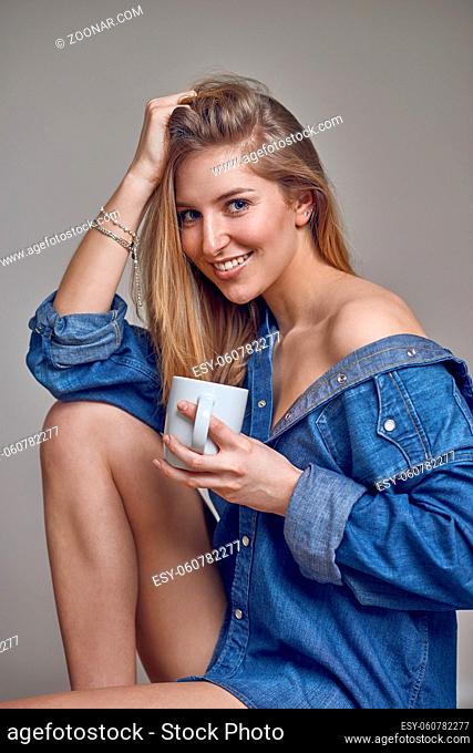 Attractive trendy young blond woman in denim shirt opened over the shoulder and bare legs sitting holding a mug of coffee smiling happily at camera with her...