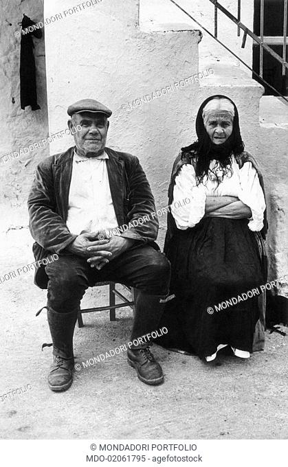 An old Italian couple in commoner clothes; they are sitting side by side in a little court in the historic center of a rural village