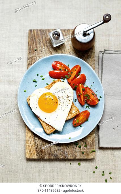 A slice of toast topped with a heart-shaped fried egg served with cherry tomatoes