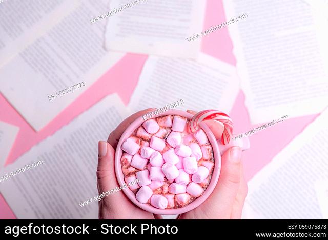 Woman holding in her hands a cup of hot chocolate with pink mini marshmallows and an Xmas candy cane, over a table with book pages