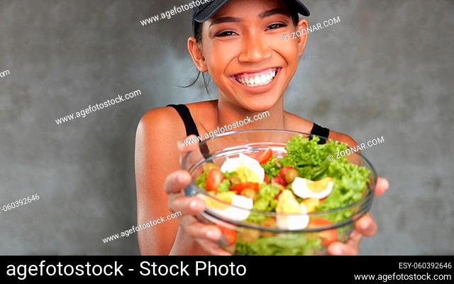 Portrait of beautiful young smiling woman in black t-shirt holding bowl with healthy salad over concrete background