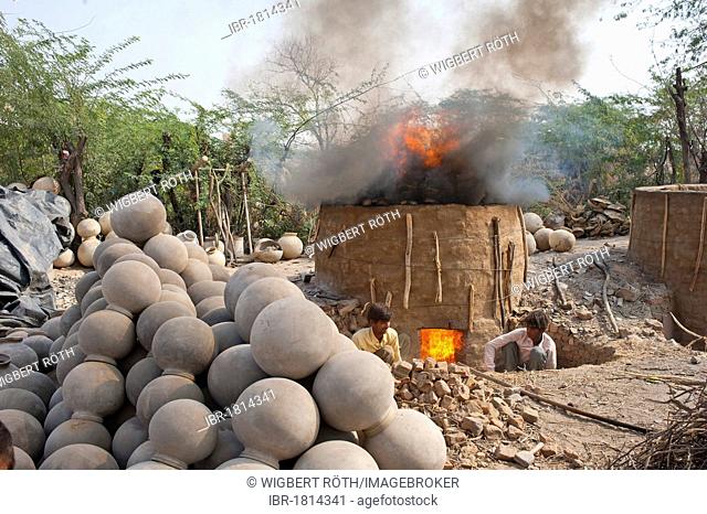 A clay kiln for firing clay water jugs, two potters puting more firewood on the kiln, in front stacked finished water jugs, Bishnoi, Jodhpur, Rajasthan, India