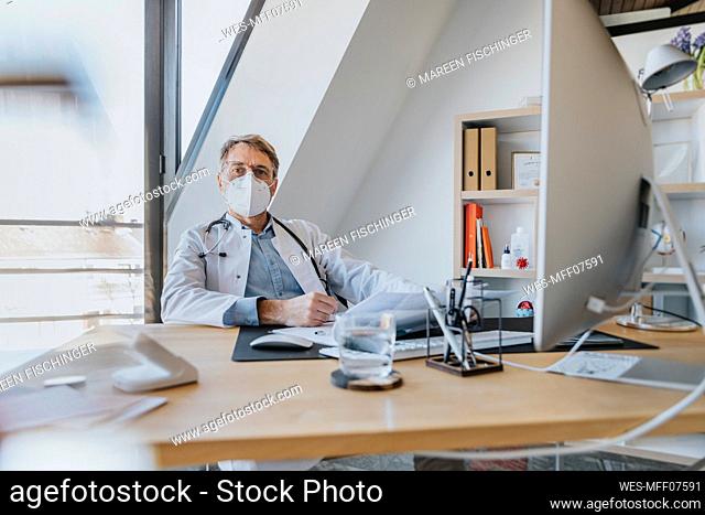 Mature doctor wearing protective face mask sitting by desk at doctor's office
