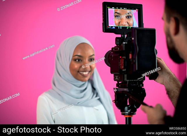 male videographer in digital studio recording video on professional camera by shooting female muslim woman wearing hijab scarf plastic pink background