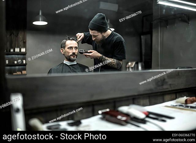 Peerless barber with a beard and a tattoo is cutting the hair of his bearded client in the barbershop. He is using a cutting comb and a hair clipper
