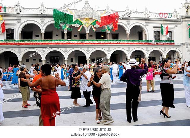 Couples dancing in the Zocalo with facade of government buildings behind hung with bright decorations in the national colours for Independence Day celebrations