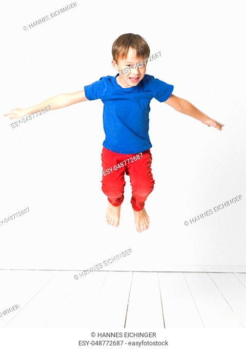 cool, young and cute boy with blue shirt and red trousers is jumping high in the studio in front of white background and white wooden floor