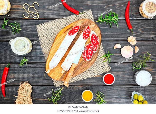 Sliced cured sausage with spices and a sprig of rosemary on dark wooden rustic background