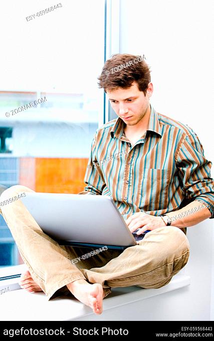 Young casual office worker sitting at office window working on laptop computer