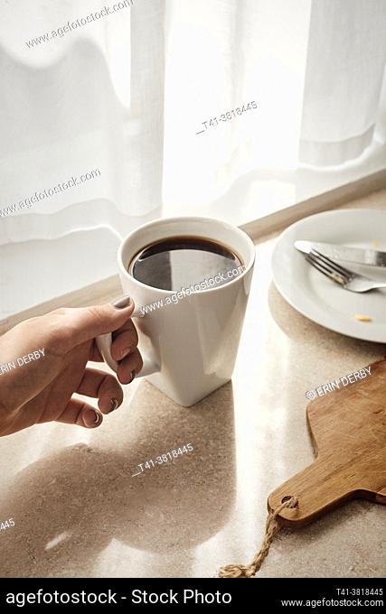 A woman's hand holding a white mug with coffee in a sunstreaked kitchen