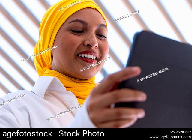 Happy biracial businesswoman with hijab using tablet outside office