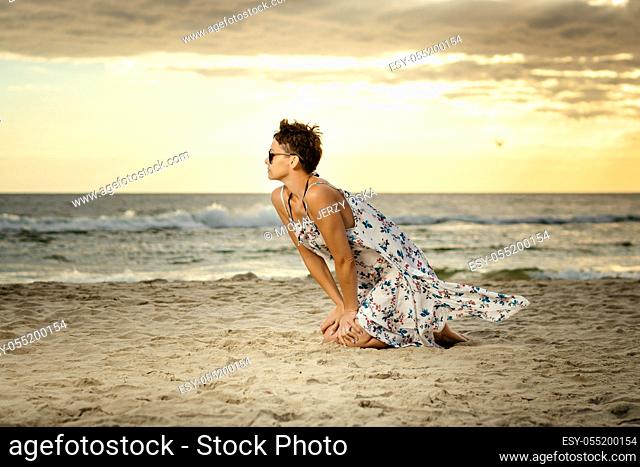 beautiful woman in a long dress sitting on the sand at the beach at sunset