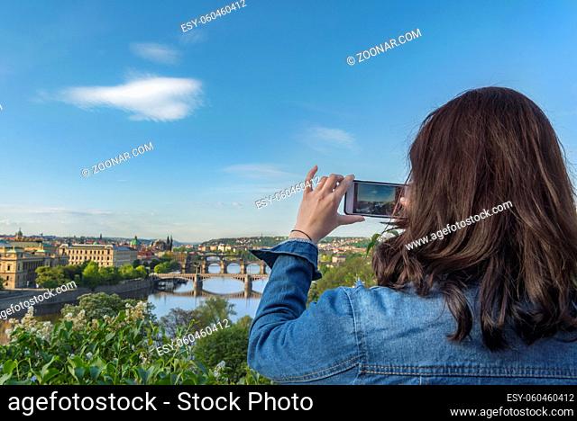 Brunette woman, from behind, taking photos with her smartphone with the Vltava river, its bridges, and the Prague city, in the Czech Republic