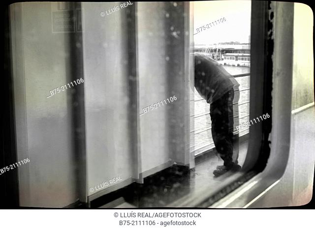Reflection in a window of a passenger boat of a man leaning on the railing of the deck overlooking the sea