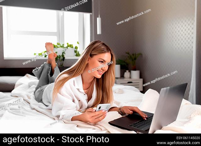 Pretty smiling Caucasian girl lays on bed in bedroom and use laptop for remote work. Communication, technology, remote work concepts in gray interior apartment