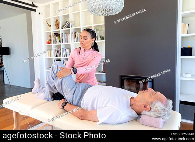 Female biracial physiotherapist stretching caucasian senior man's leg lying on table at home