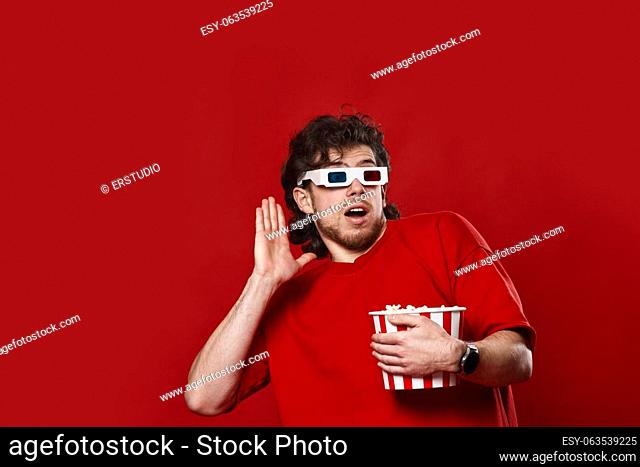 scared handsome man in 3d-glasses holding bucket of popcorn on red background