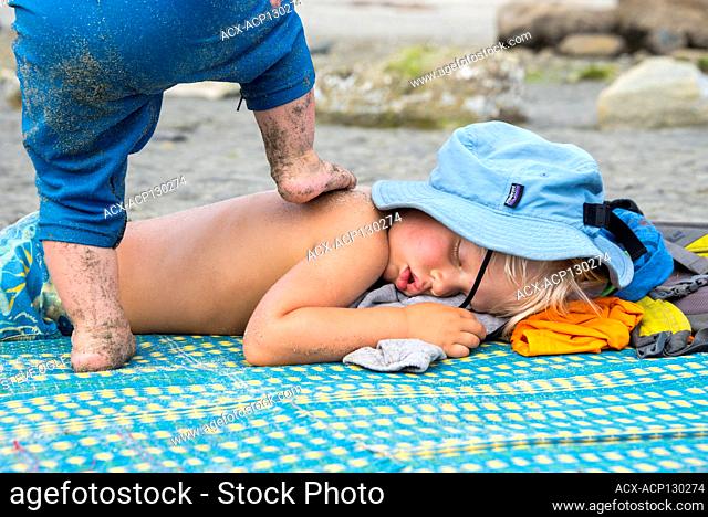 A toddler walks on his sleeping brother on a beach blanket