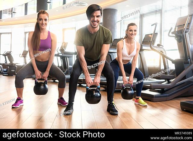Three fit young people smiling and looking at camera while exercising kettlebell swings during full body workout at the gym