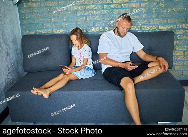 Father and daughter browsing mobile phone while relaxing together on sofa at home, woman and girl play on smartphones while resting on couch on weekend day