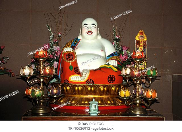 White porcelain statue of the Maitreya Bodhisattva or Laughing Buddha seated on a golden lotus flower with coloured lamps and flowers at the Vihara...