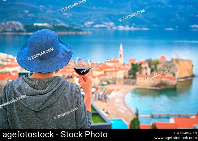 Caucasian woman wearing blue hat standing in an apartment balcony and holding glass with red wine looking at the Budva town below, Montenegro